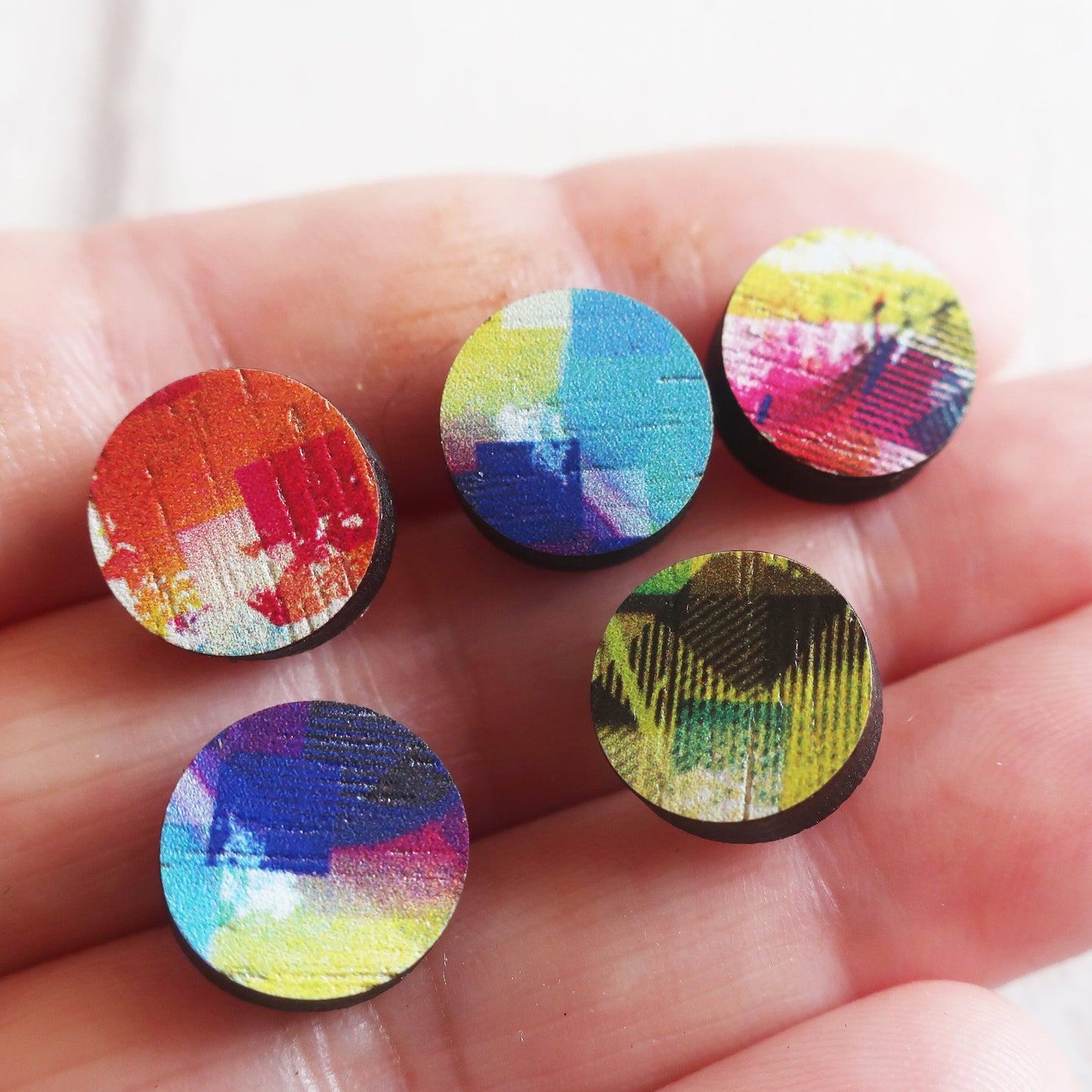 Five colorful round wooden earrings held in a hand against a white background