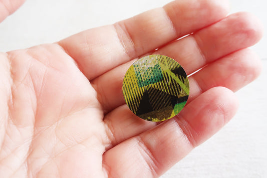Round pin badge with green and black pattern held in a hand against a white background