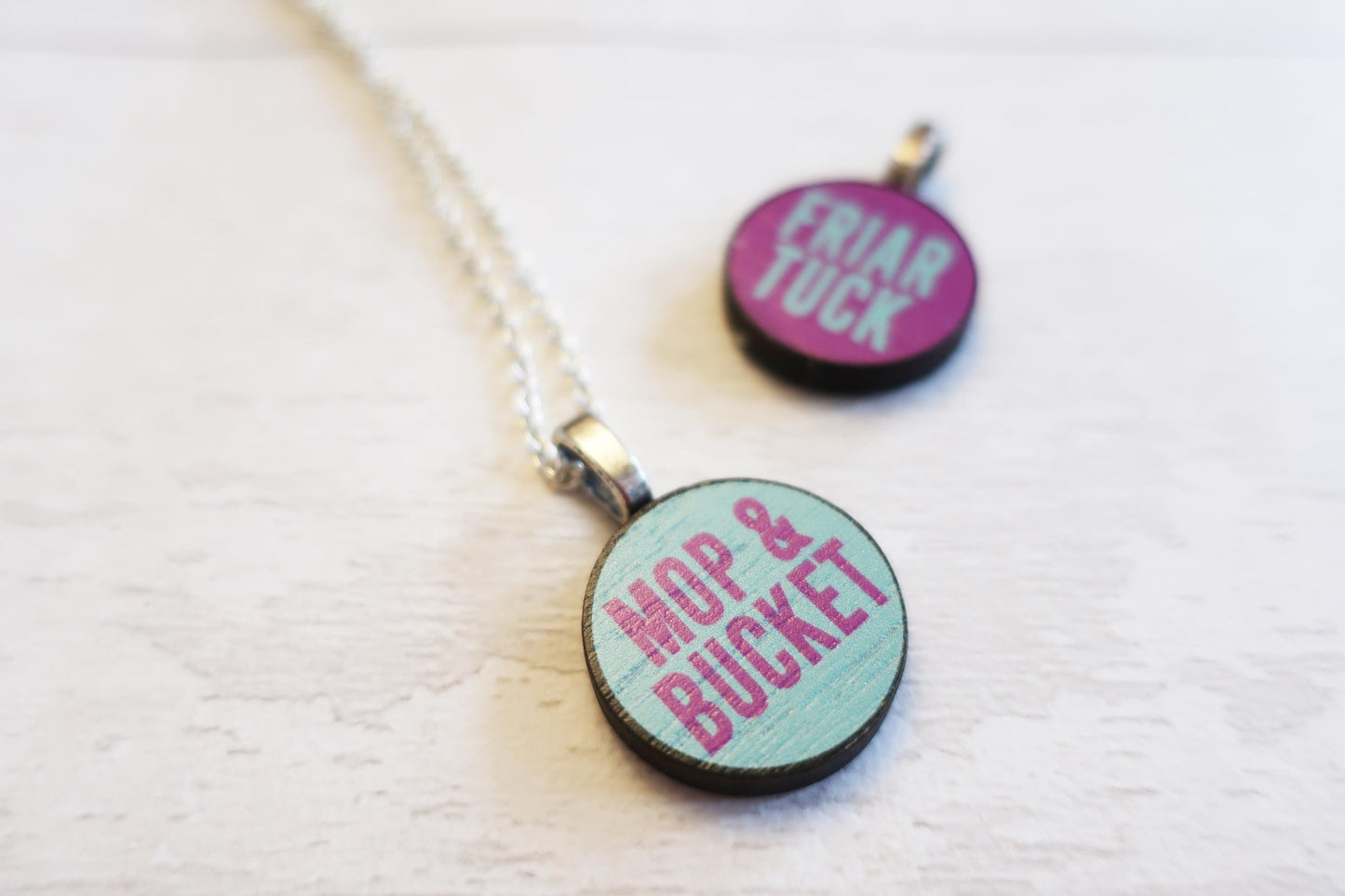 Two round pendants on a chain with text 'FRIAR LUCK' and 'MOP & BUCKET' on a light background.