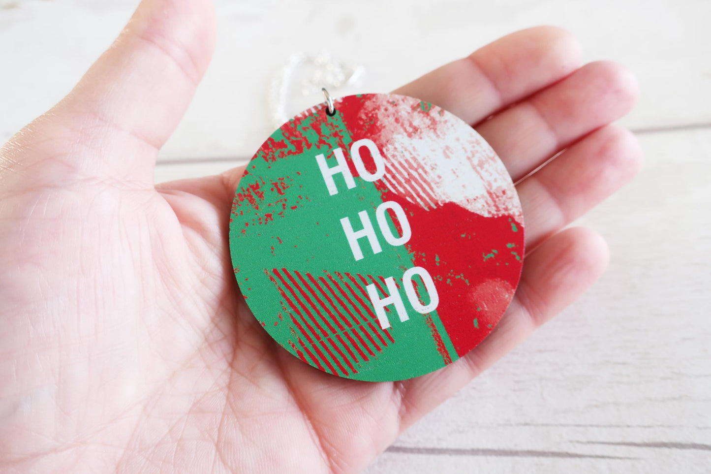 Hand holding a round, red and green necklace with 'HO HO HO' text on a light wooden surface.