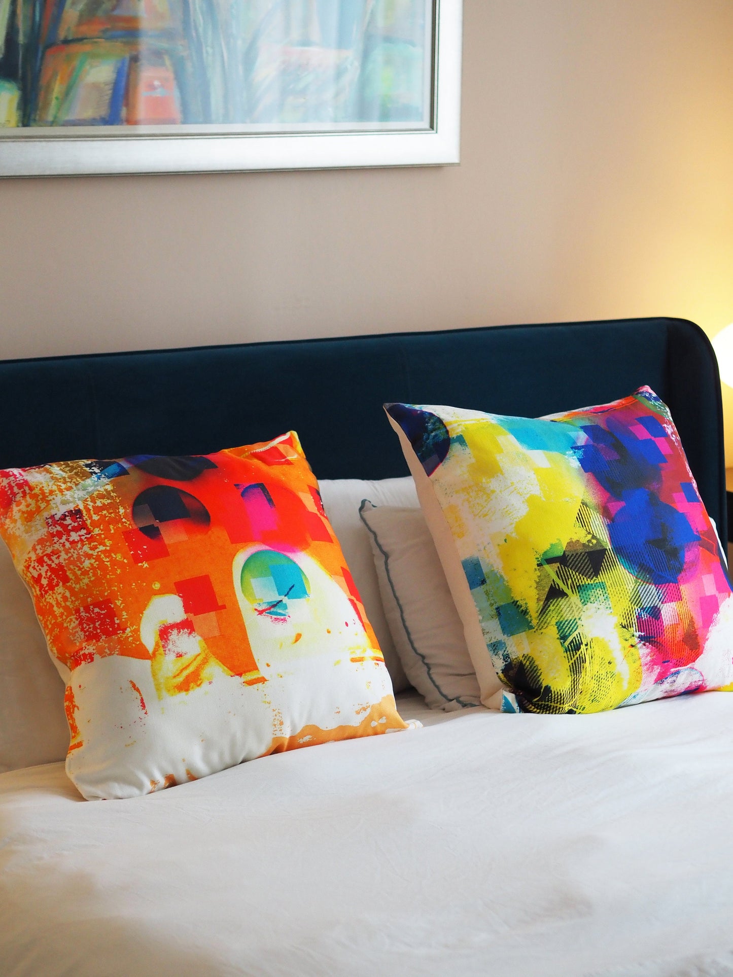 Colorful abstract-patterned pillows on a bed with a framed picture in the background.