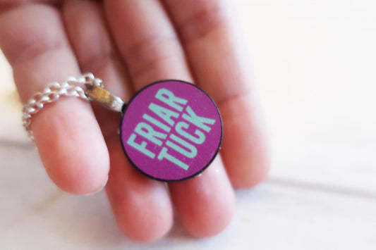 Round purple pendant with 'FRIAR TUCK' text held between fingers on a light background