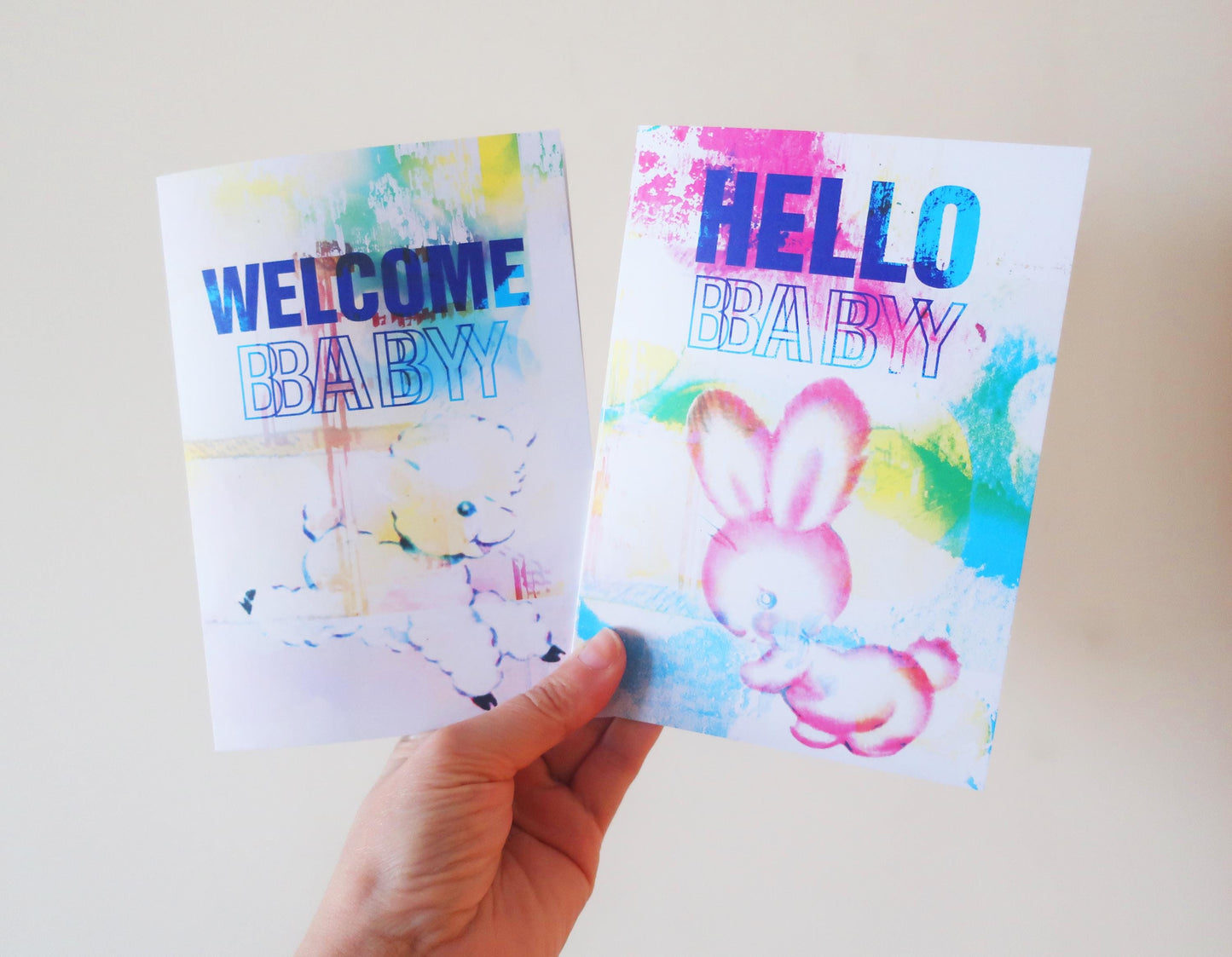 Two colorful greeting cards with 'Welcome Baby' and 'Hello Baby' text held by a hand against a light background.