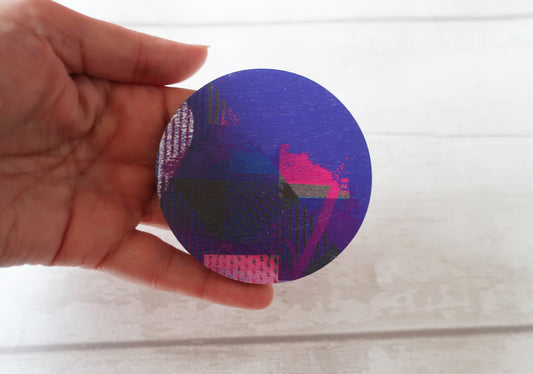 Hand holding a round, purple brooch with geometric patterns on a white background