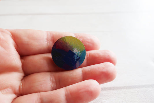 Hand holding a round, multicolored pin badge against a white background