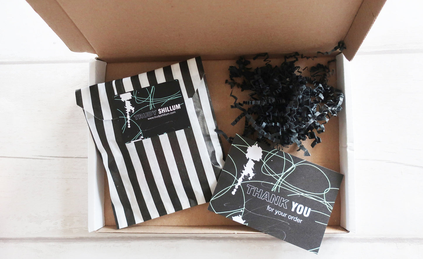 Open cardboard box with striped package, black decorative items, and a 'Thank You' card on a white wooden surface.