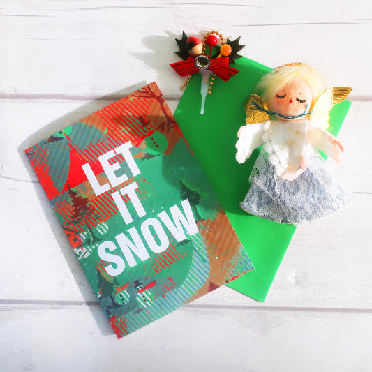 Christmas-themed card with 'LET IT SNOW' text and a small angel figurine on a white wooden surface.