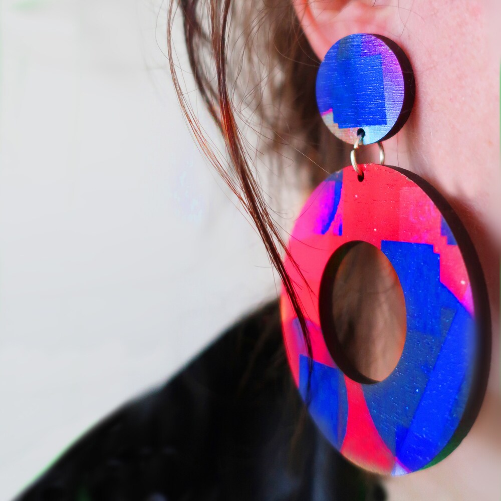 A large pair of circular hoop earrings in pink and purple being modelled. The ear and some hair is in shot.