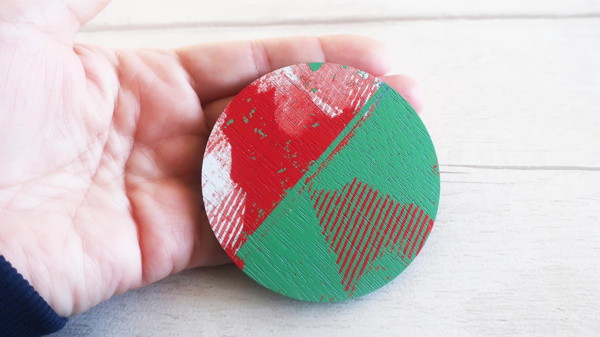 Round red and green brooch held by a hand on a light wooden surface