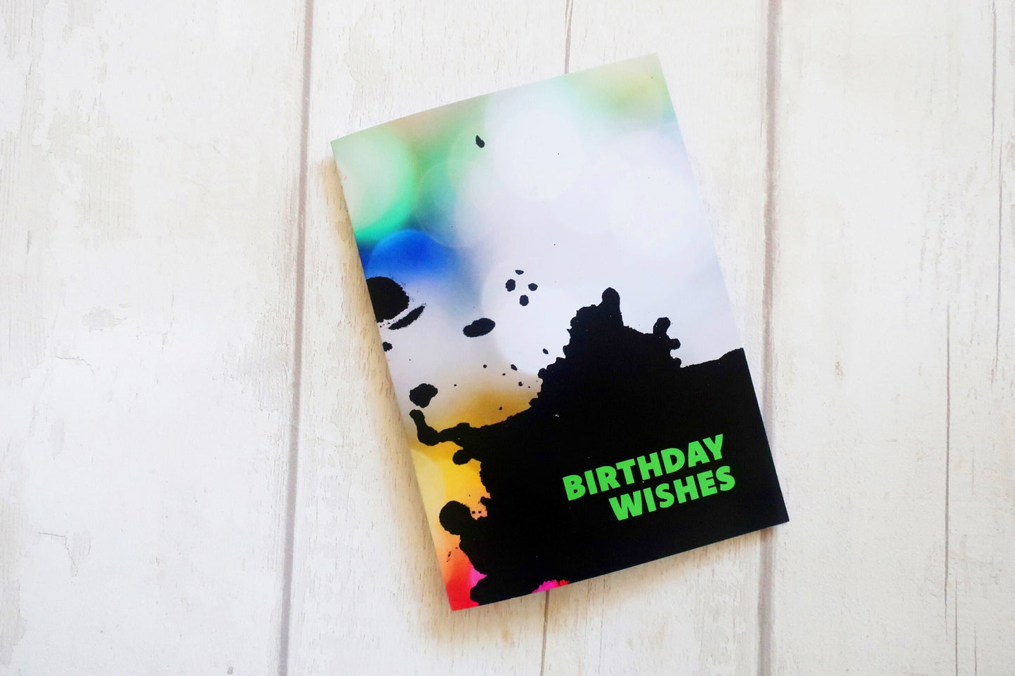 Greeting card with colorful splatter design and 'Birthday Wishes' text on a white wooden surface