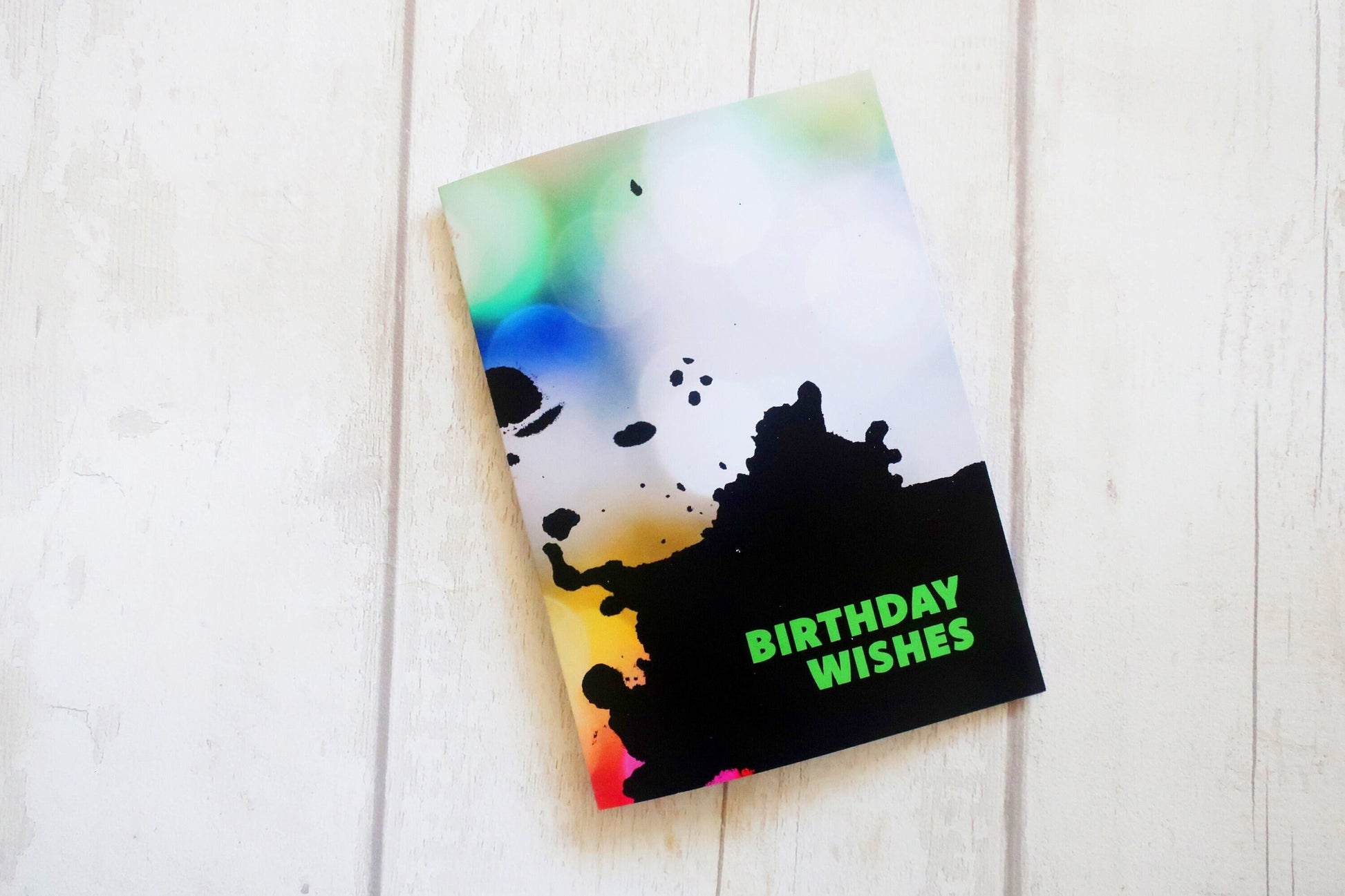 Greeting card with colorful splatter design and 'Birthday Wishes' text on a white wooden surface