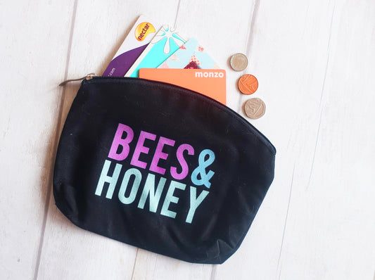 Black pouch with 'BEES & HONEY' text, containing bank cards and coins on a light wooden surface.