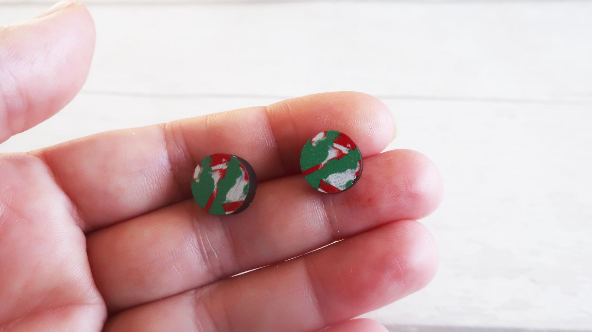 Small round earrings with green and red design held between fingers against a white background