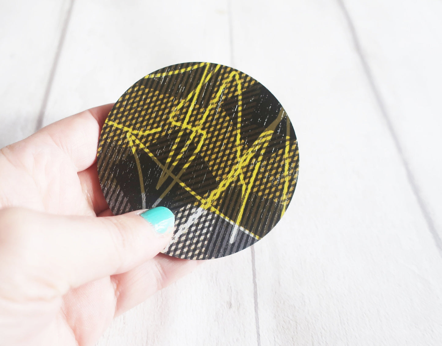 Round black and yellow patterned brooch held by a hand with light blue nail polish on a white background.