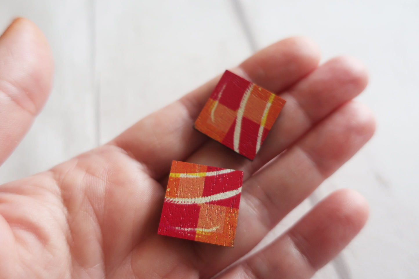 Two small square earrings with red and orange patterns held between fingers against a white background.