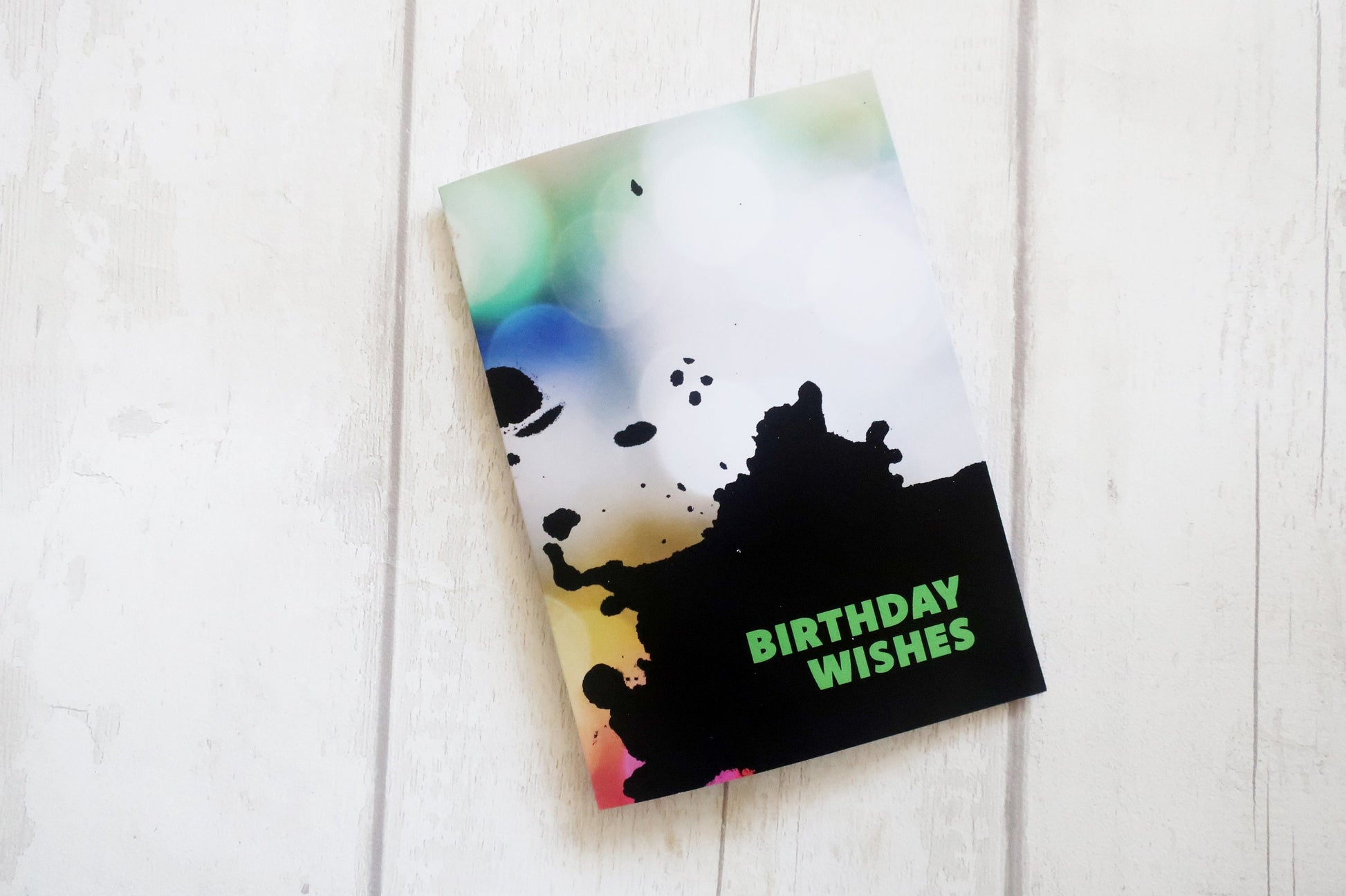 Greeting card with colorful splatter design and 'Birthday Wishes' text on a white wooden surface