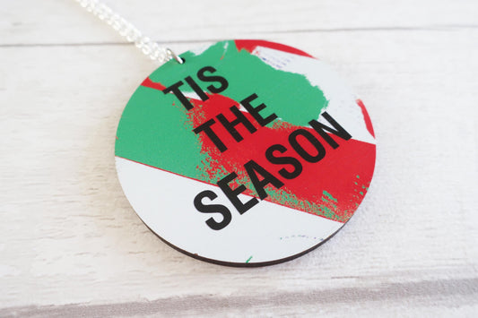 Round decorative necklace with 'TIS THE SEASON' text on a white wooden surface