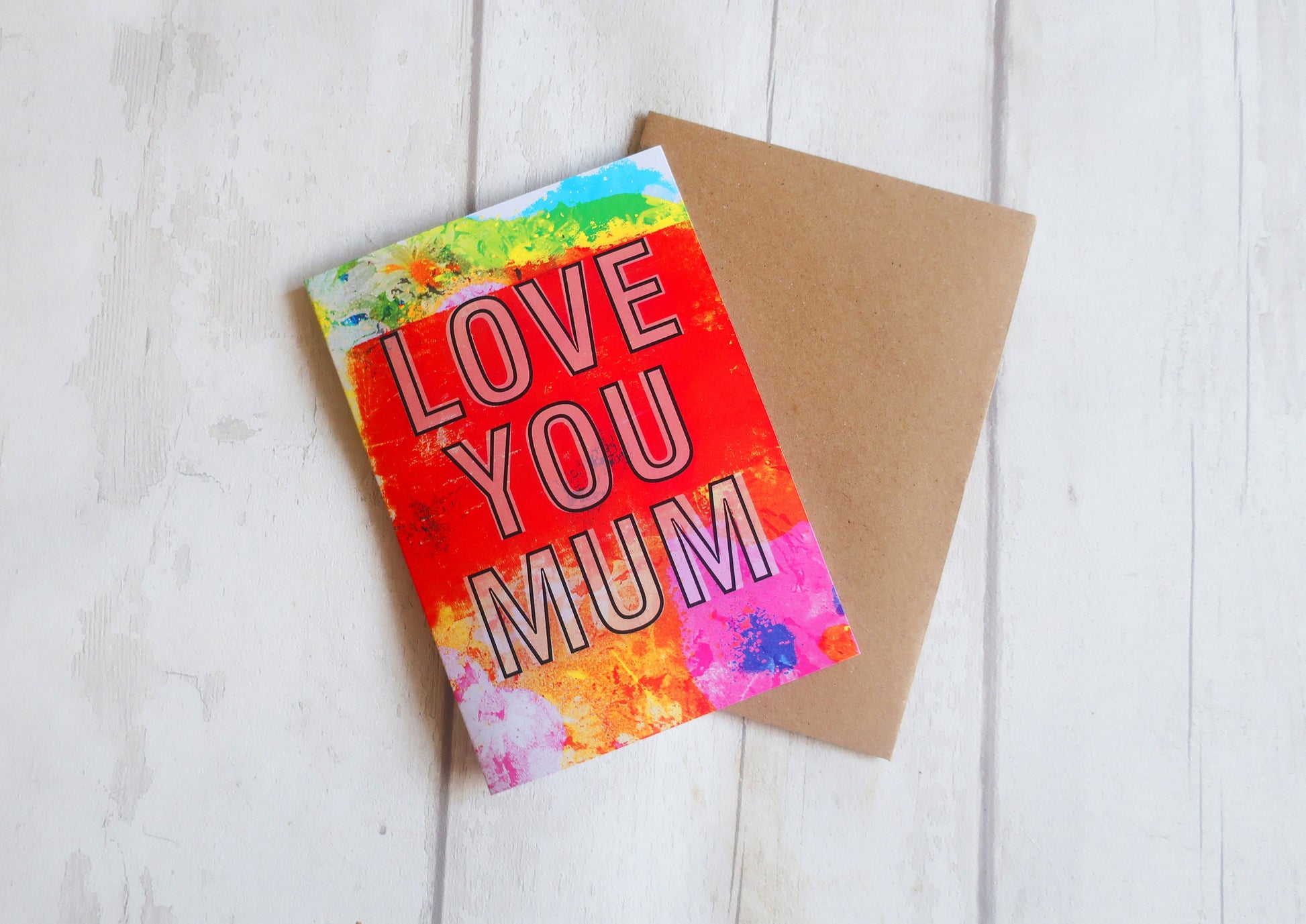 Greeting card with 'LOVE YOU MUM' text on a colorful background, placed on a wooden surface.