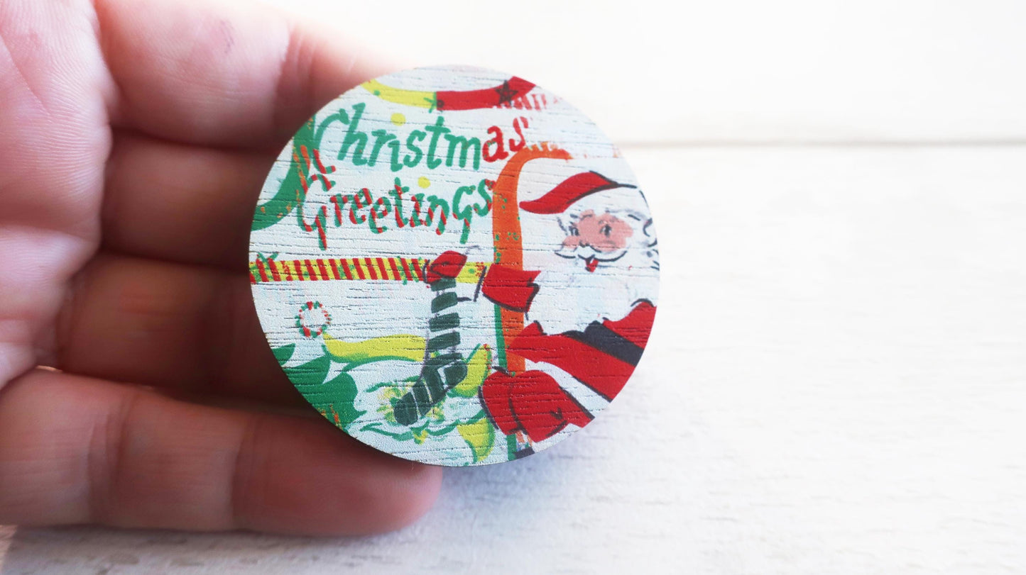 Round Christmas-themed badge with Santa Claus and 'Christmas Greetings' text held by a hand.