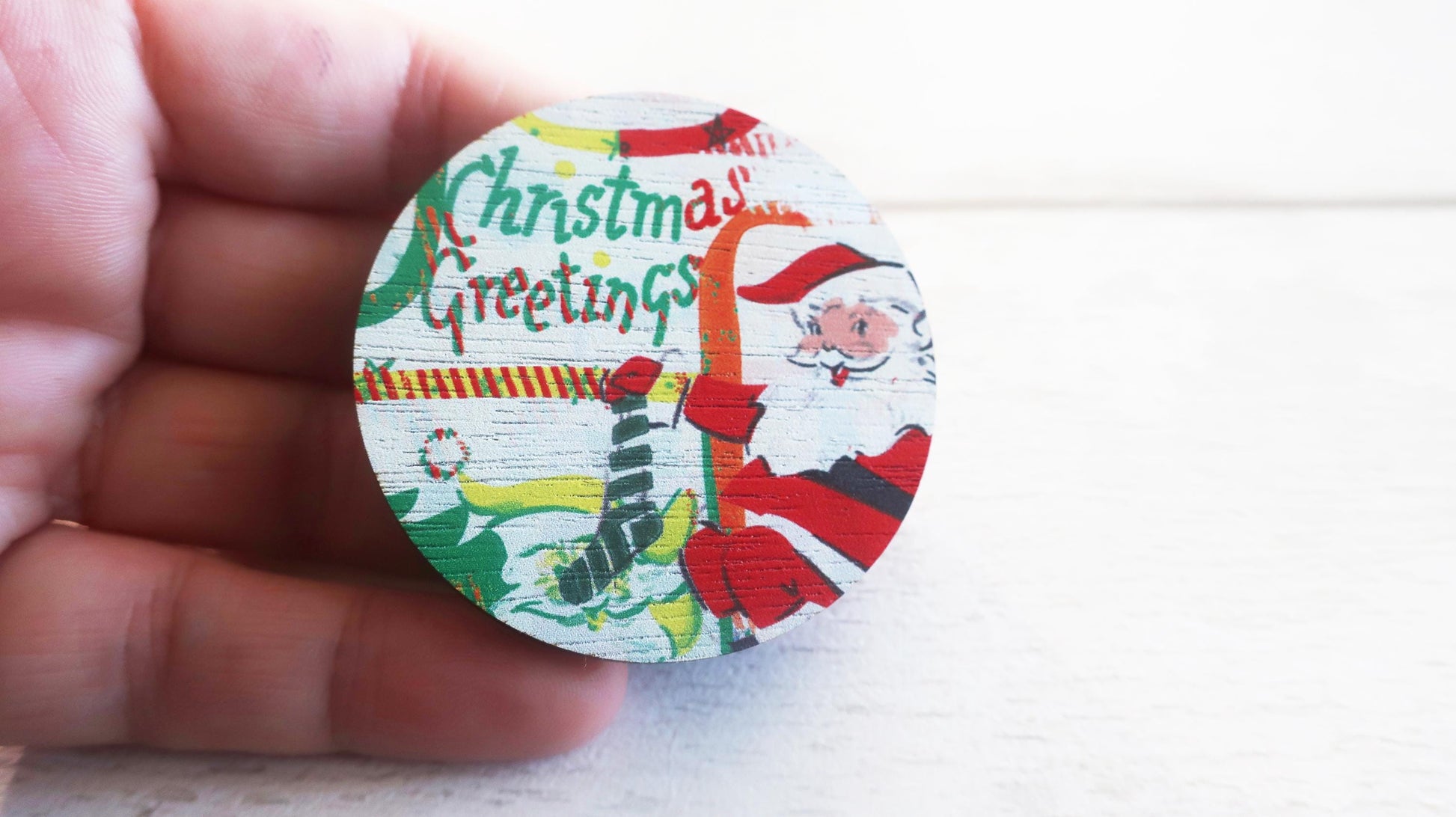 Round Christmas-themed badge with Santa Claus and 'Christmas Greetings' text held by a hand.