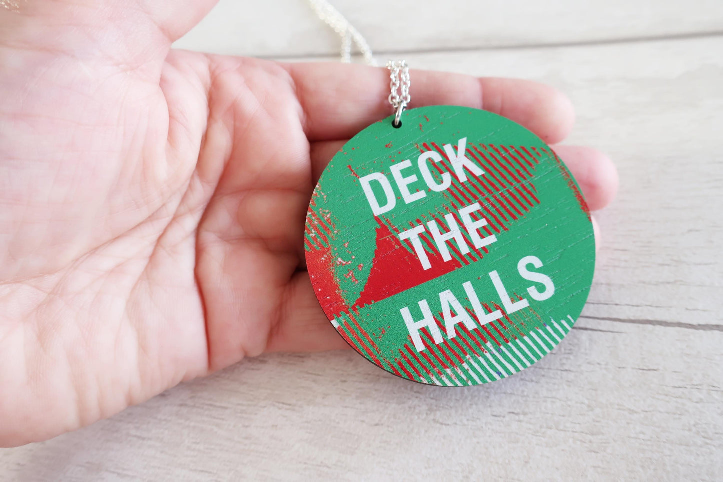 Green necklace with 'Deck the Halls' text held in a hand on a light wooden surface