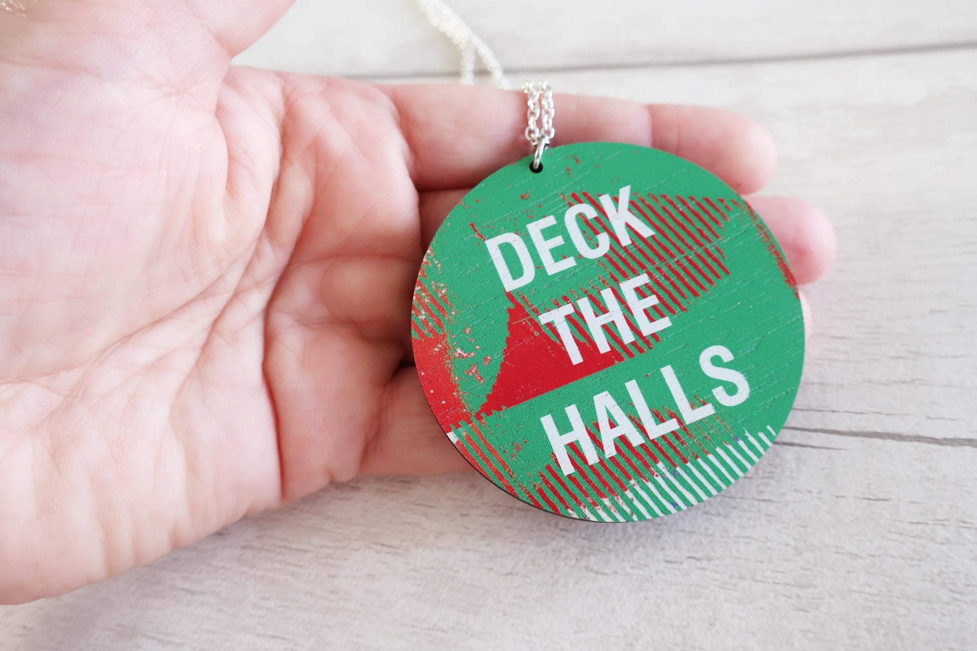 Green necklace with 'Deck the Halls' text held in a hand on a light wooden surface