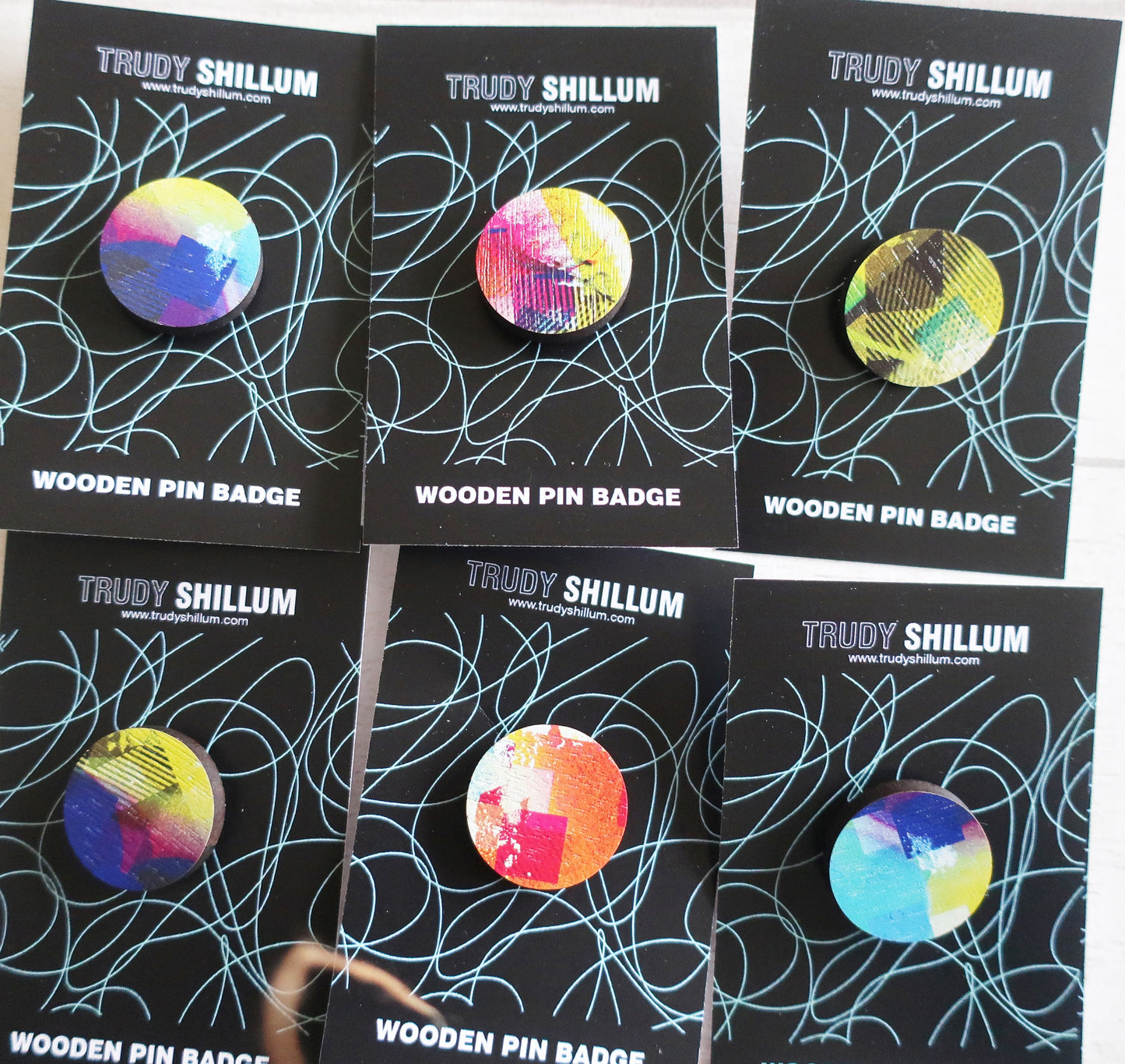 Trudy Shillum wooden pin badges on black cards with colorful designs.