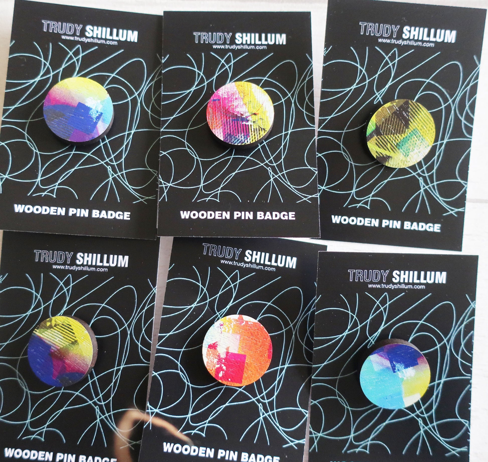 Trudy Shillum wooden pin badges on black cards with colorful designs.