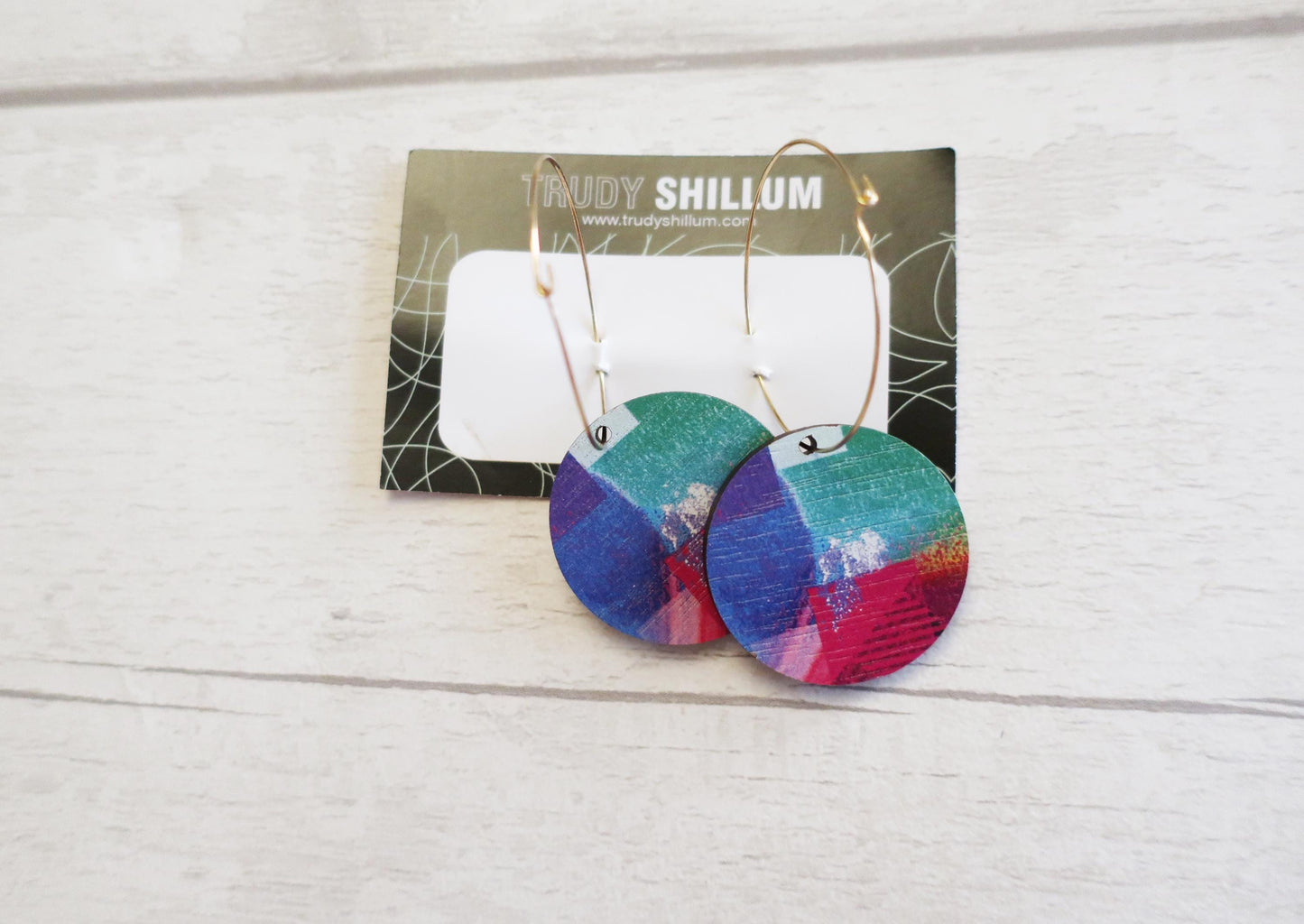 Colorful earrings on a card with 'Trudy Shillium' branding on a wooden surface.