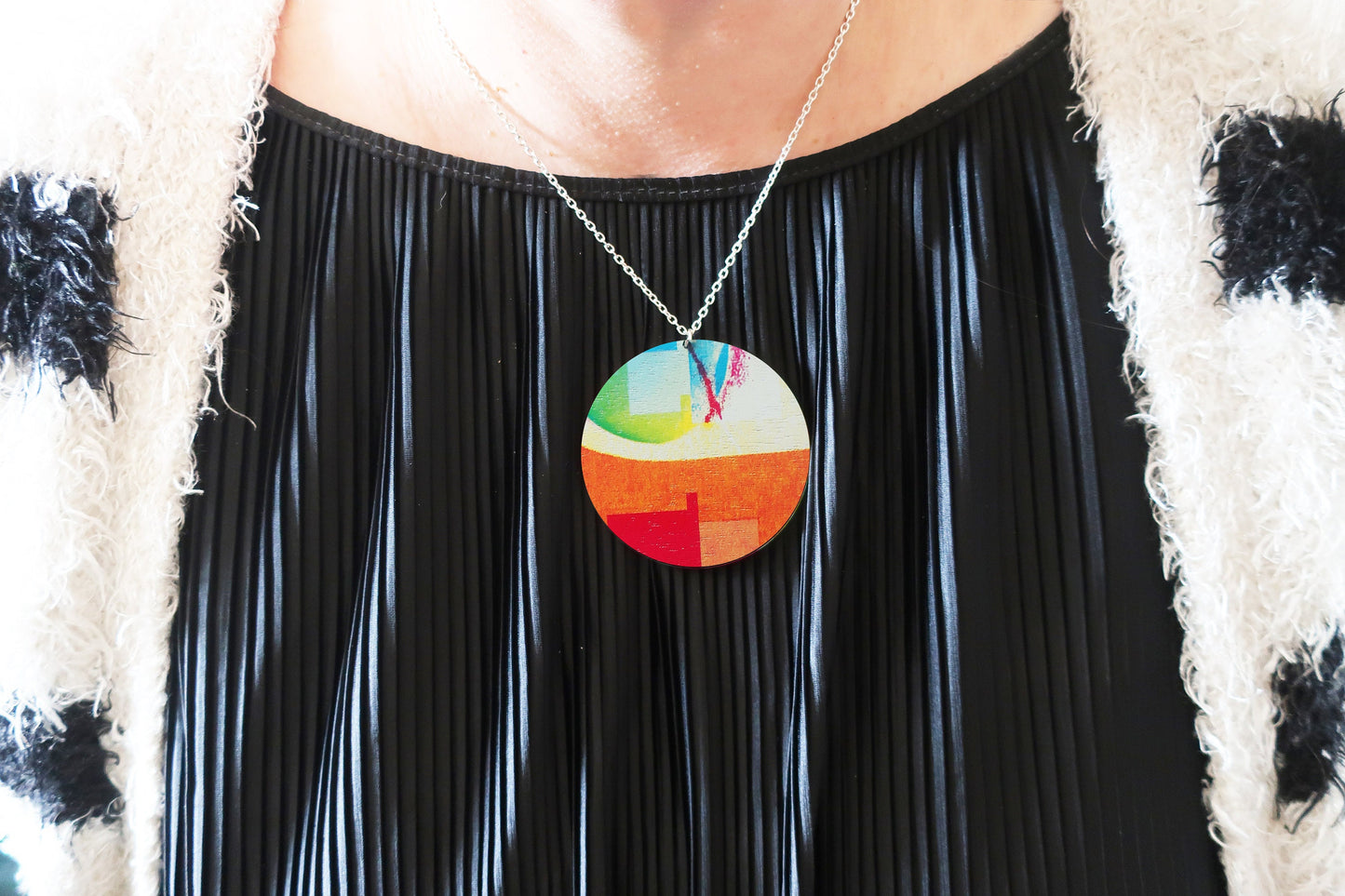 Person wearing a colorful pendant necklace with a black pleated dress and patterned sweater.
