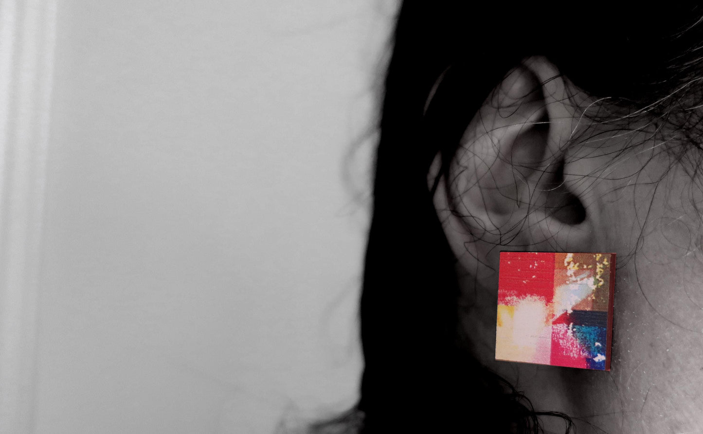 Close-up of an ear wearing a colorful square earring against a neutral background