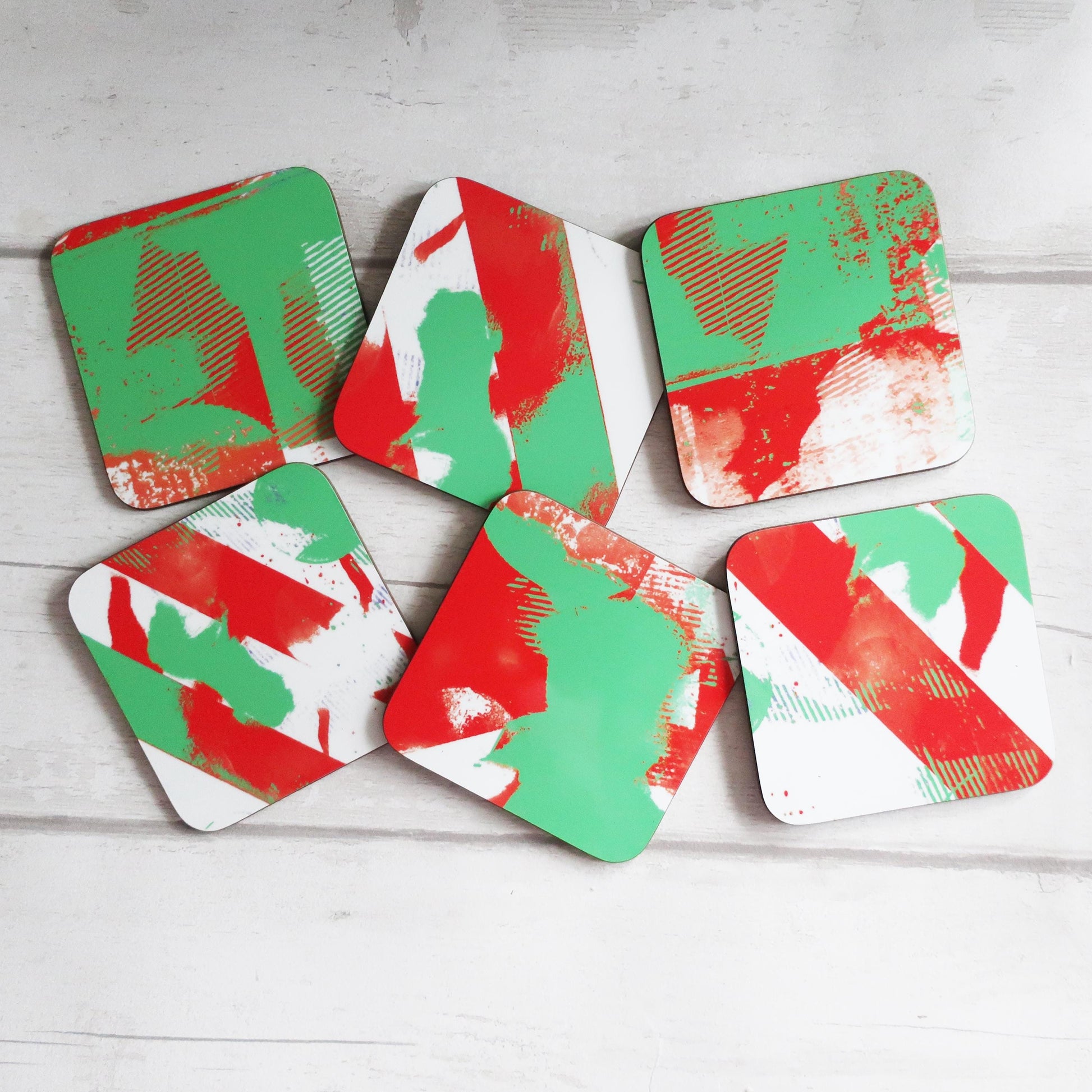 Set of six geometric coasters with red, green, and white patterns on a light wooden surface.
