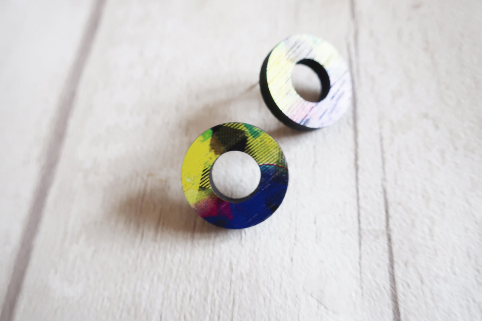Two circular earrings with colorful patterns on a light wooden surface.