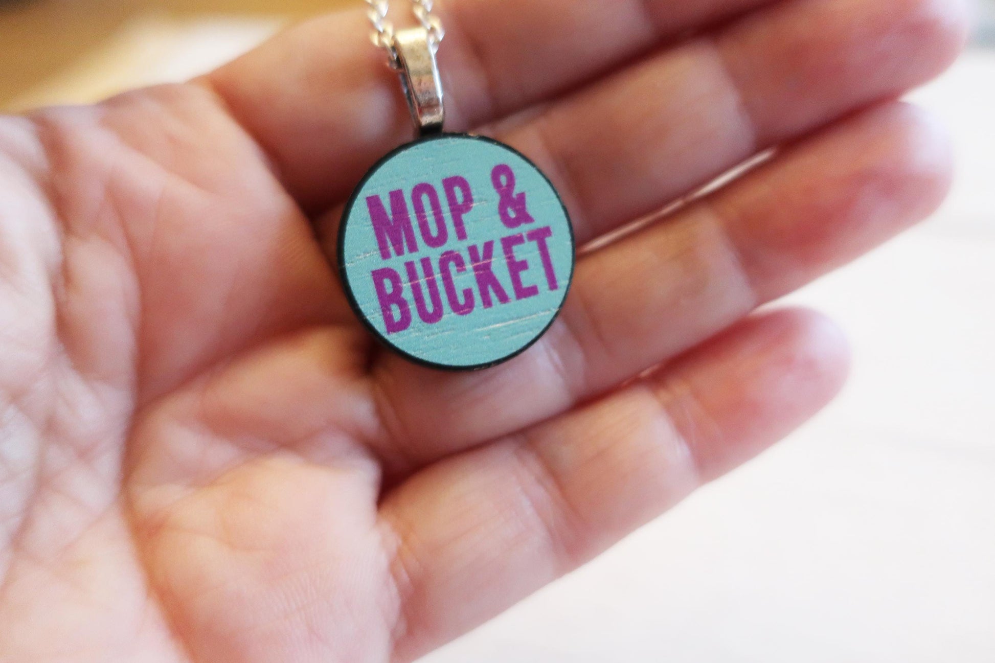 Necklace with a pendant labeled 'Mop & Bucket' held in a hand.