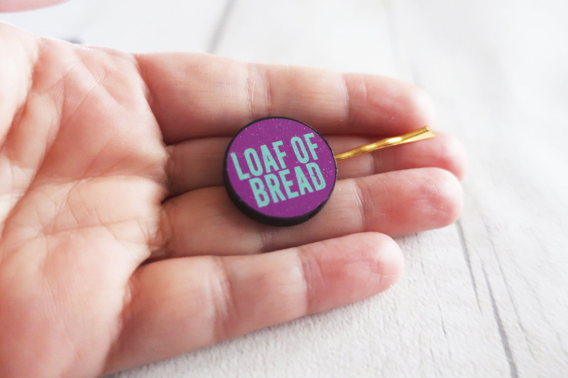 Round purple hairclip  with 'LOAF OF BREAD' text held in a hand on a light background