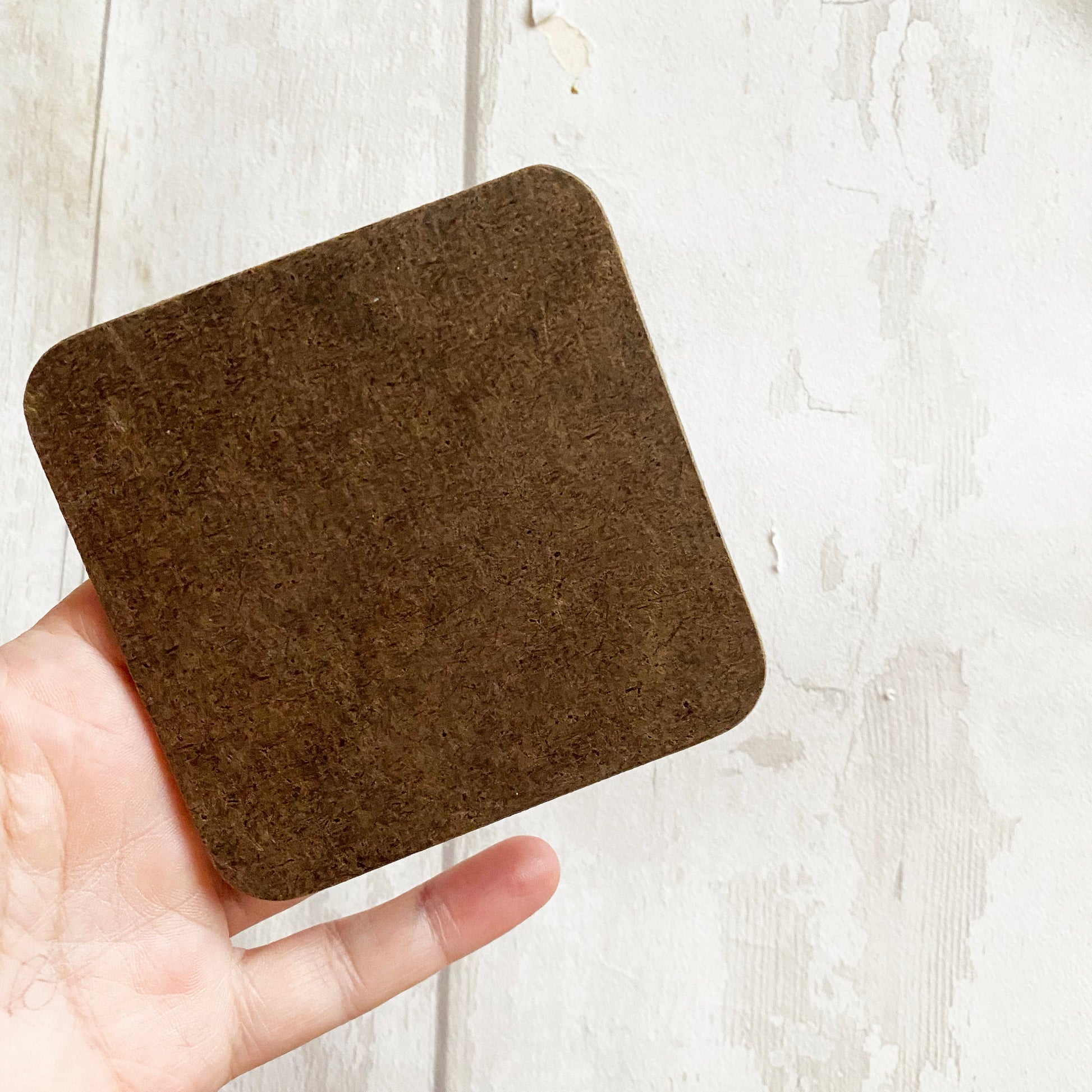 Brown square coaster held by a hand on a light wooden surface
