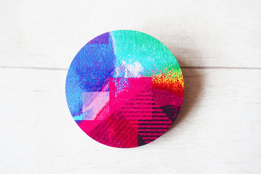 Colorful circular brooch with a gradient of blue, green, pink, and orange on a white wooden surface.