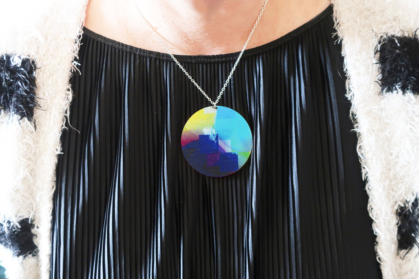 Person wearing a colorful pendant necklace with a black pleated dress and patterned sweater.