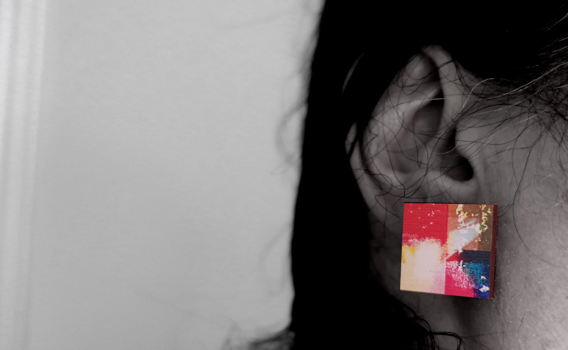 Close-up of an ear wearing a colorful square earring against a neutral background