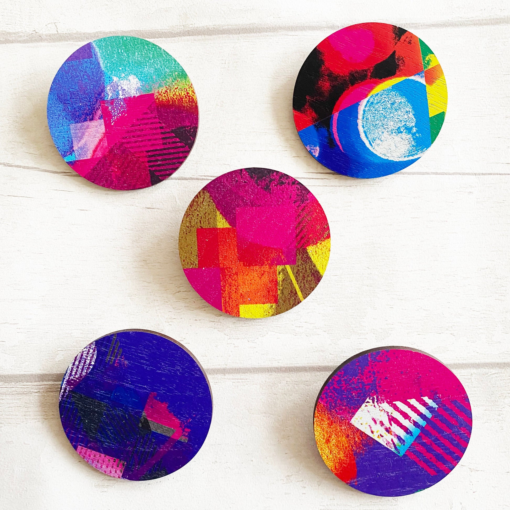 Five colorful circular brooches with abstract patterns on a white wooden surface.
