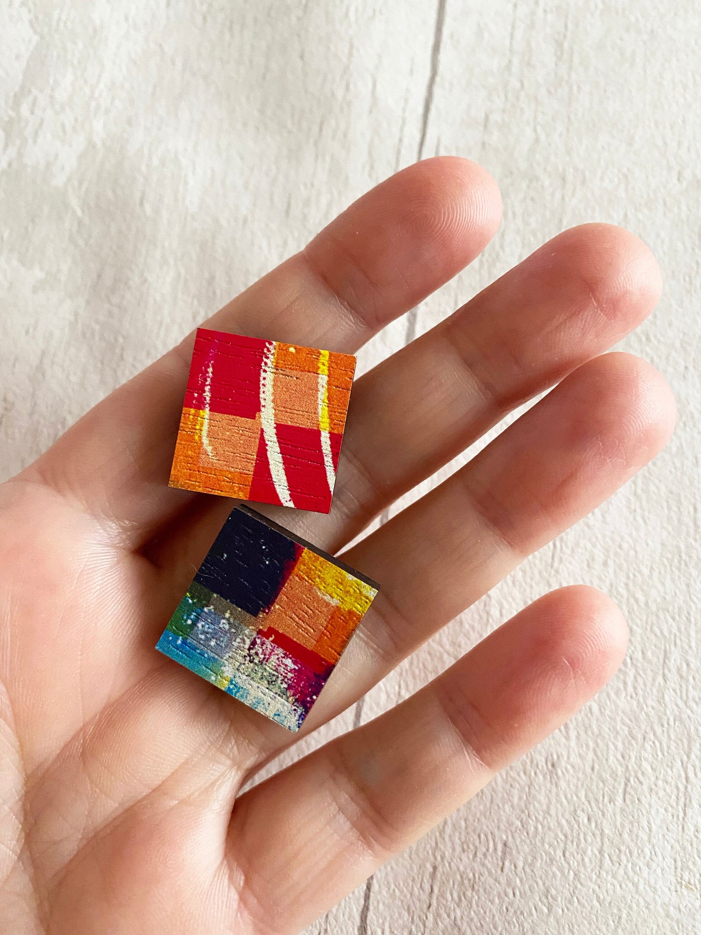 Colorful square earrings held between fingers against a light background