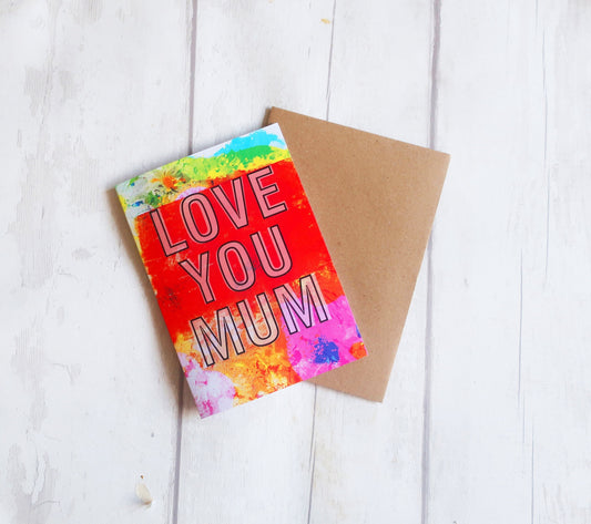 Greeting card with 'LOVE YOU MUM' text on a colorful background, placed on a wooden surface.