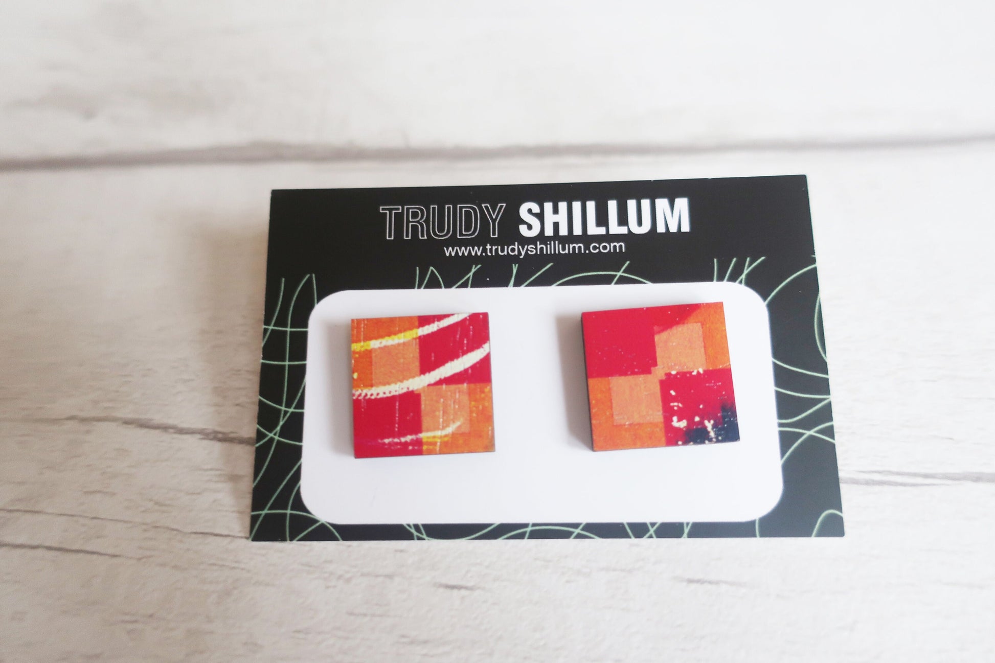 Colorful square earrings on a Trudy Shillum branded card against a light wooden background