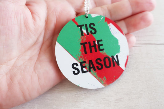 Round decorative necklace  with 'TIS THE SEASON' text on a green, red, and white background held by a hand.