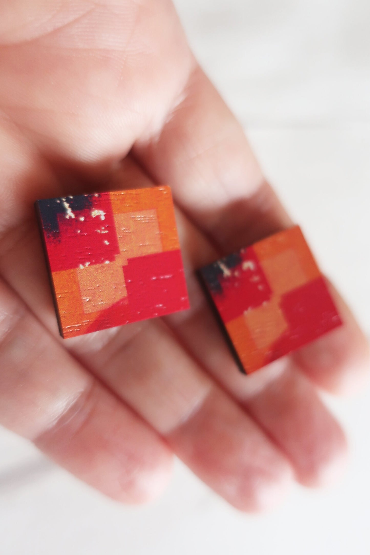 Two small square earrings with a geometric pattern held between fingers.