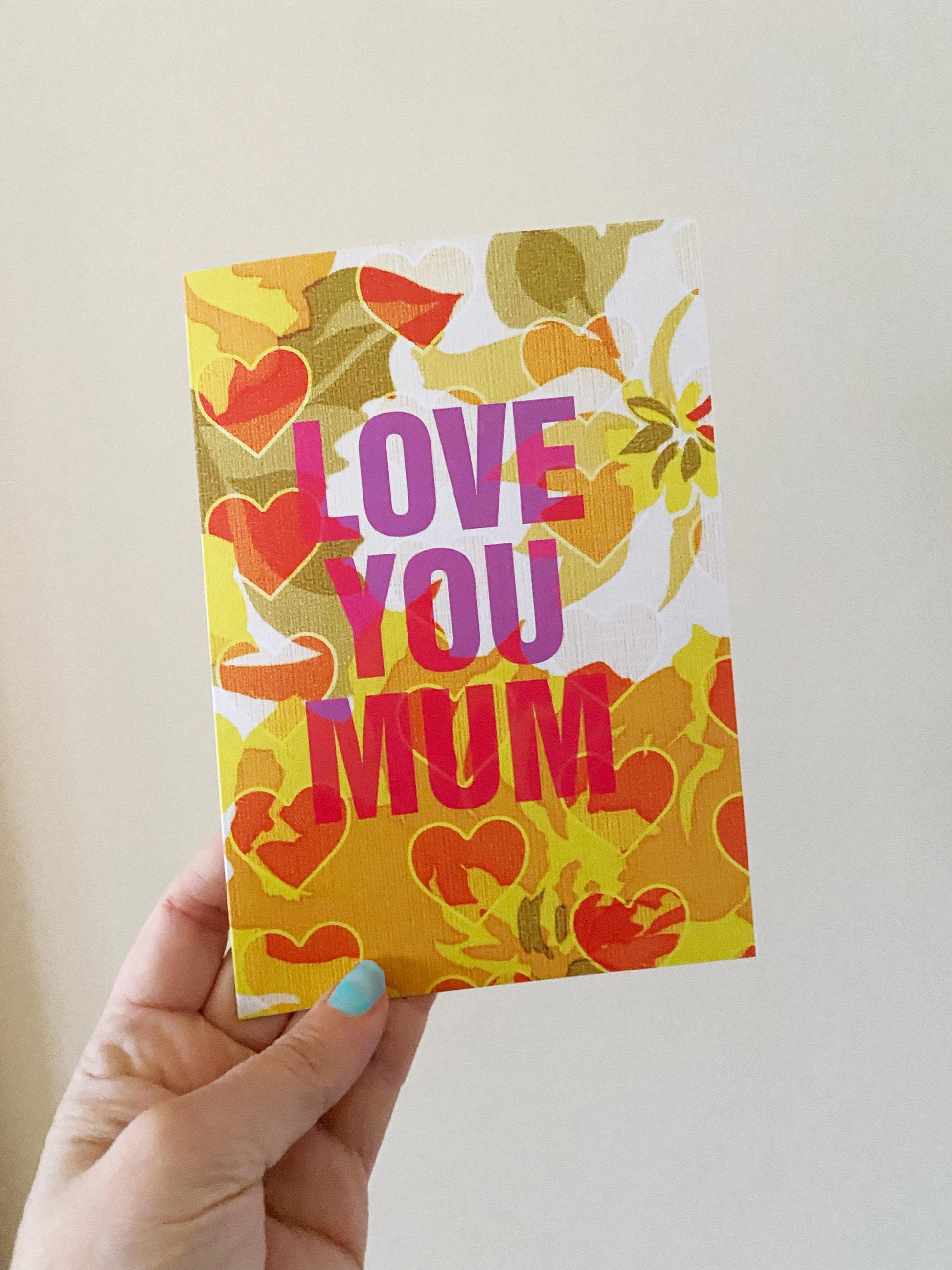 Hand holding a colorful card with 'LOVE YOU MUM' text against a plain background