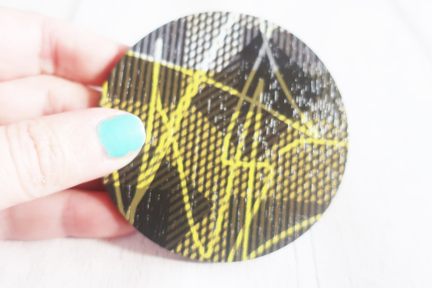 Round brooch with black and yellow pattern held by a hand with teal nail polish.