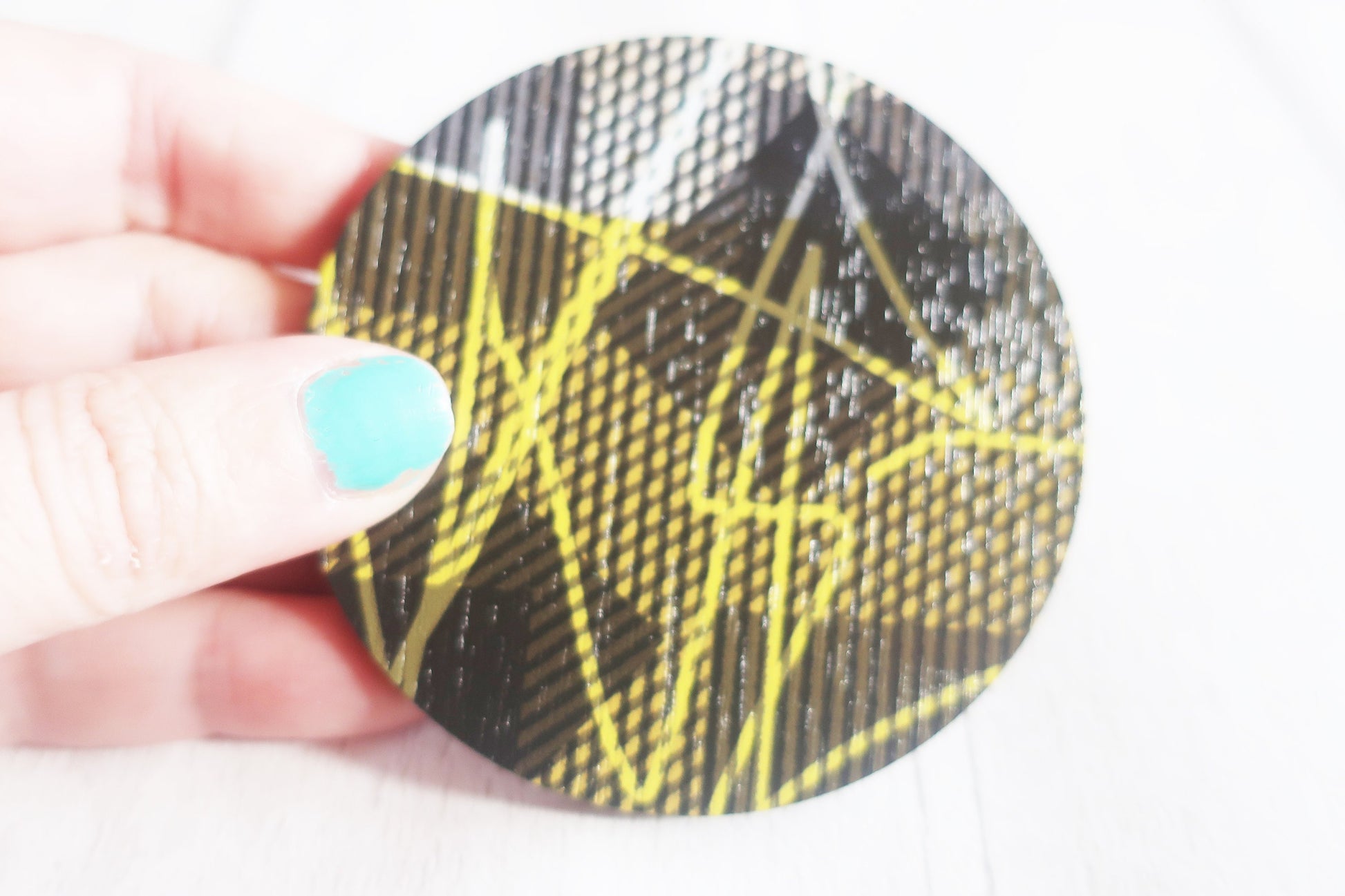 Round brooch with black and yellow pattern held by a hand with teal nail polish.
