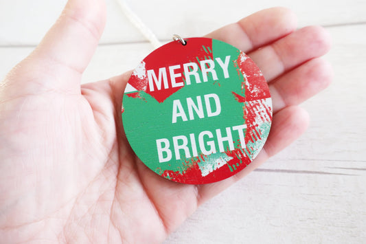Hand holding a round, red and green necklace with 'Merry and Bright' text on a white background
