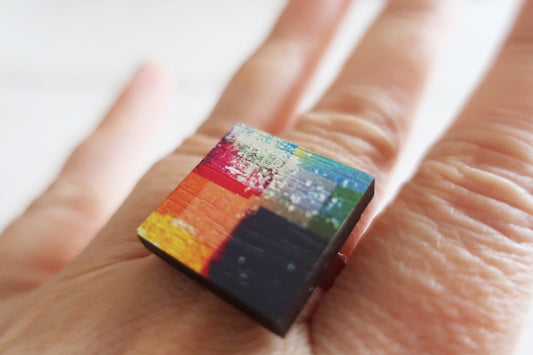 Small, colorful square ring held between fingers against a white background